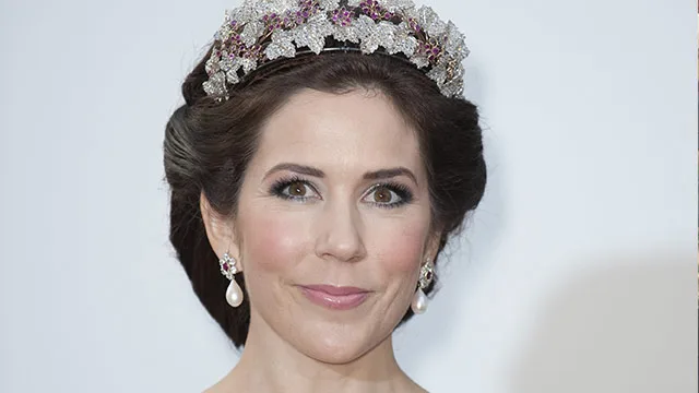 A woman with styled hair wears an ornate tiara and pearl earrings, smiling slightly against a neutral background.