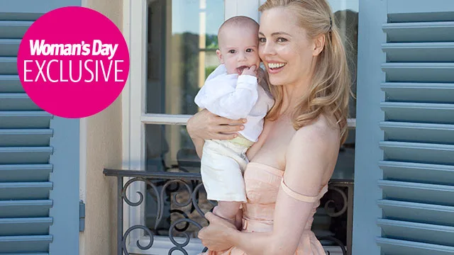A woman smiling, holding a baby in front of a building with blue shutters, "Woman's Day Exclusive" logo in corner.