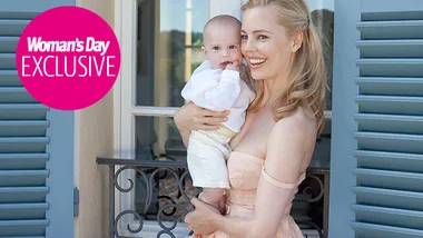 A woman smiling, holding a baby in front of a building with blue shutters, "Woman's Day Exclusive" logo in corner.