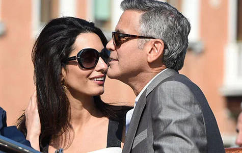 A couple smiling and leaning close to each other while wearing sunglasses, with a blurred building in the background.