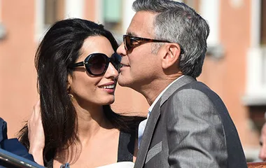 A couple smiling and leaning close to each other while wearing sunglasses, with a blurred building in the background.