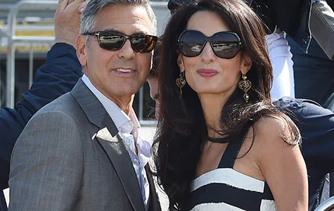 Couple in sunglasses smiling outdoors, she wears striped dress and earrings, he wears grey suit.