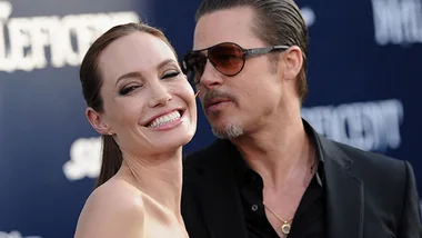 "Two people smiling on a red carpet event, one wearing sunglasses and the other with a bright smile."