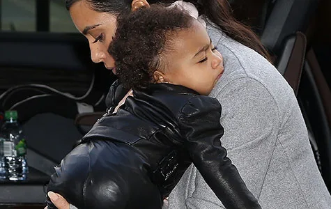 A woman carrying a sleeping child in a black leather outfit, outside a vehicle with a water bottle visible.
