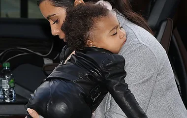 A woman carrying a sleeping child in a black leather outfit, outside a vehicle with a water bottle visible.