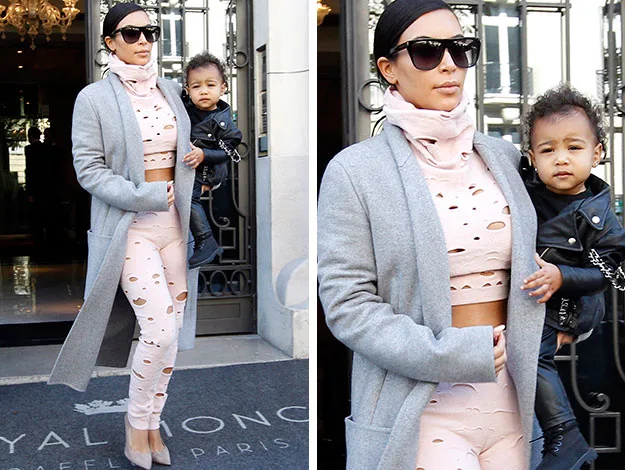 Woman in gray coat and sunglasses carrying a young child in black jacket, exiting a building with glass doors.