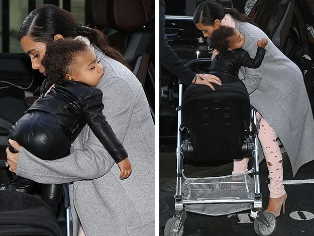 A woman in a gray coat carrying a sleeping child in a black jacket and pants, beside a stroller.