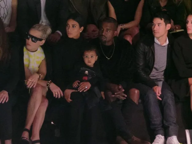 Group of adults seated at a fashion event, including one holding a child, with focused expressions and diverse styles.