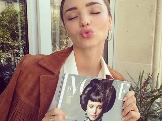 A woman in a brown jacket puckers her lips holding a Vogue magazine issue with model cover.
