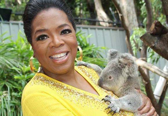 A smiling person in a yellow shirt holds a koala, with lush greenery in the background.
