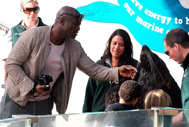 Man interacting with a seal, surrounded by others and onlookers at a marine exhibit.
