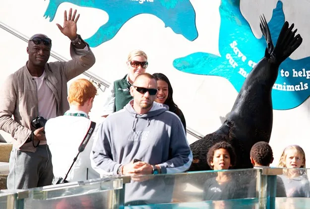 Group of people at a zoo, including a man waving and children watching a sea lion perform against a blue mural backdrop.
