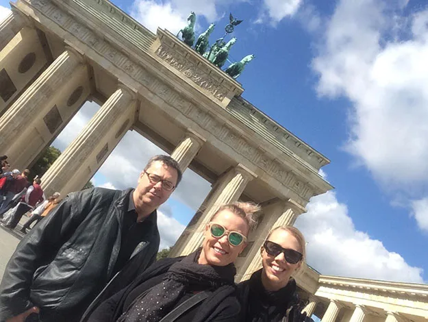 Three people smiling in front of the Brandenburg Gate in Berlin under a blue, cloudy sky.