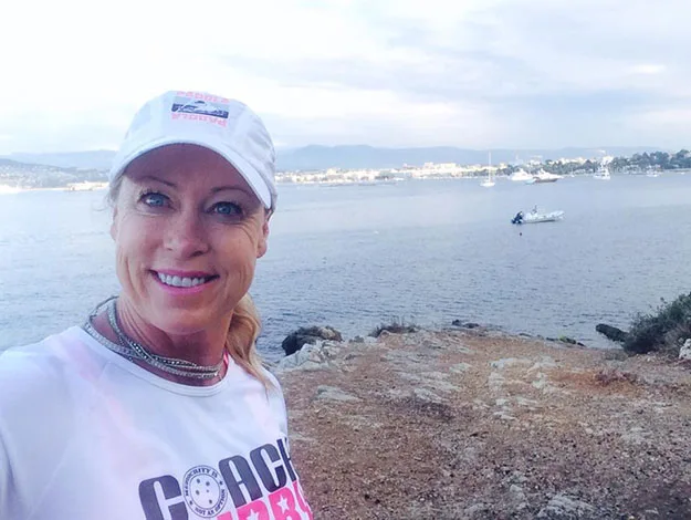 Woman in a "Coach" shirt and cap smiling by the sea with boats and distant shoreline in the background.