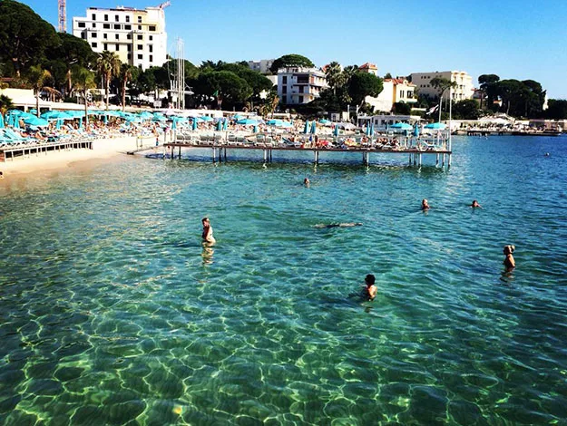 Beach with clear turquoise water, swimmers, and sunbathers on loungers, buildings in the background under a blue sky.