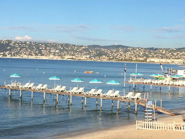 Pier with white lounge chairs and turquoise umbrellas over calm water; distant hills and clear blue sky in background.