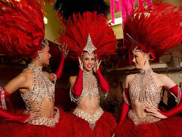Showgirls in elaborate red feathered costumes and sparkling headpieces get ready backstage, smiling and chatting.