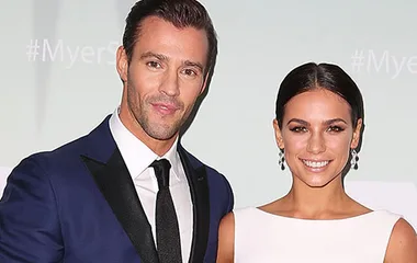 A man in a dark suit and a woman in a white dress smiling, standing together against a light background.