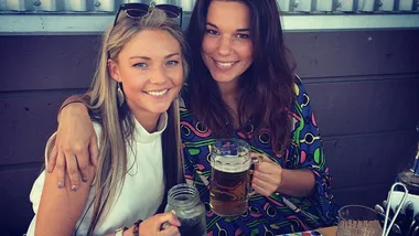 Two women smiling, sitting together at a table. One holds a mason jar, the other a mug of beer, enjoying a sunny day.
