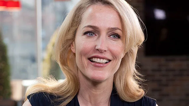 Gillian Anderson smiling, with long blonde hair, in an indoor setting with a blurred background.