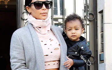 Mother in sunglasses holds toddler in black leather jacket, standing outside a building with metal gates.