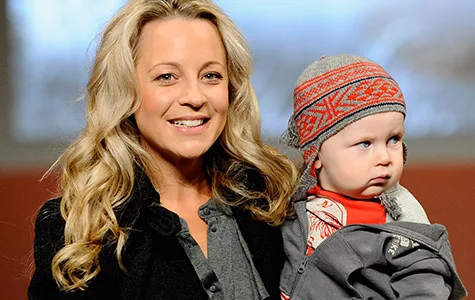 Carrie Bickmore smiling, holding a baby in a winter outfit with a patterned beanie, indoors with a blurred background.