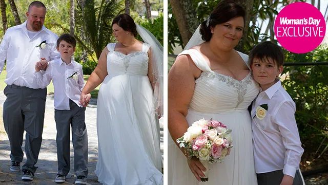 A young boy walks a smiling bride down a tropical aisle, holding hands, with a man beside them; close-up with them embracing.