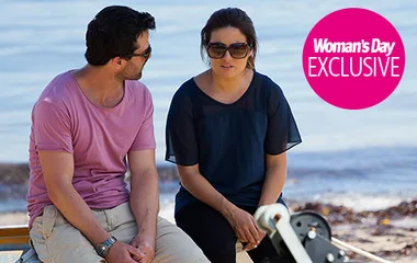 Two people sit on a beach, engaged in conversation. The woman wears sunglasses, and there's a "Woman's Day Exclusive" logo.