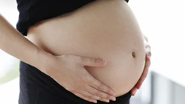 Pregnant woman holding her belly, wearing a black shirt, with a window background.
