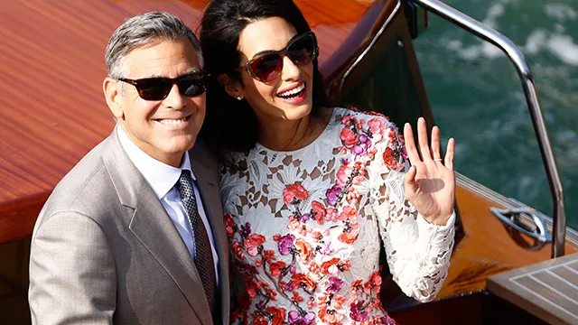 Two people smiling on a wooden boat, the woman waving. She wears a floral dress; he wears a grey suit.