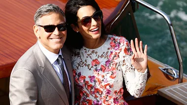 Two people smiling on a wooden boat, the woman waving. She wears a floral dress; he wears a grey suit.