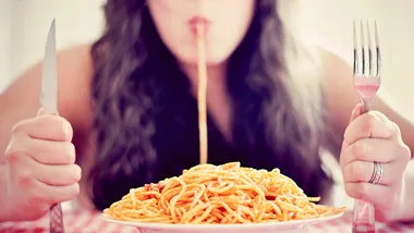 Woman eating spaghetti from a plate with a fork and knife in hands, illustrating a pasta meal setting.