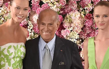 Oscar de la Renta with two models in floral dresses, standing in front of a backdrop of pink and white flowers.