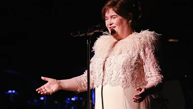 A woman in a lace dress sings passionately on stage under warm lighting, holding a microphone with outstretched arms.