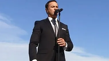 Singer performing on stage in a suit with a microphone against a clear blue sky.