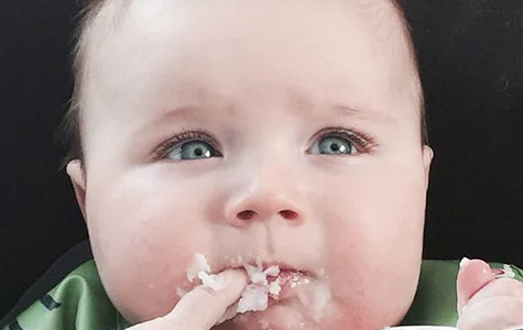 Baby touching face with messy food-covered hand, looking upward with wide eyes.