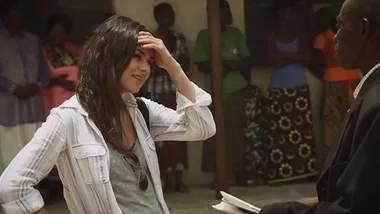 Mila Kunis, wearing a white shirt, interacts warmly with a man holding a book, surrounded by a group of people.