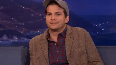 Man in a brown jacket and cap sits on a talk show set, mountain background with city lights in the distance.
