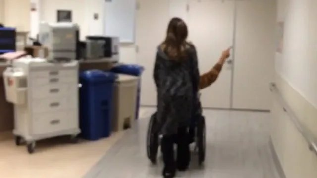 Person in wheelchair being pushed down a hospital corridor, pointing forward.