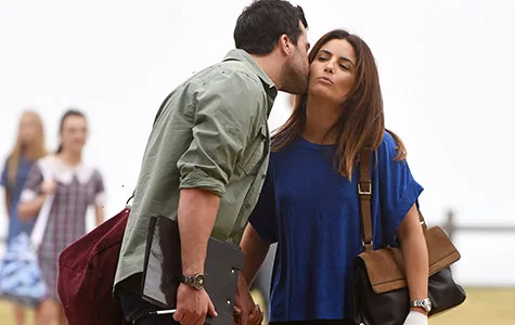A person gives Ada Nicodemou a cheek kiss on a beach, holding bags, with a blurred background of people and ocean.