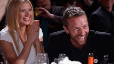 Two people smiling and clapping at a dining event, surrounded by glasses and bottles.