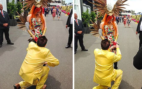 Man in yellow suit proposes to woman in colorful dress and orange headdress at outdoor event; spectators in the background.