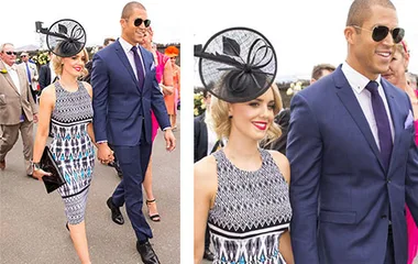 Couple in formal attire, with woman in patterned dress and hat, and man in dark blue suit, walking at Melbourne Cup event.