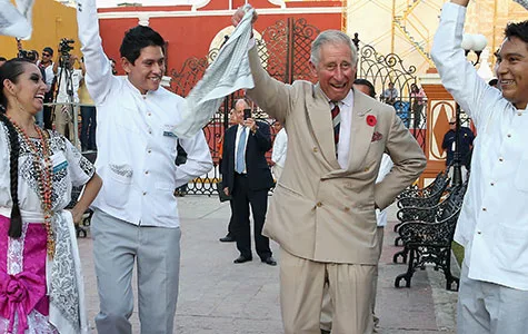 Prince Charles dancing with a group in traditional attire in Mexico, smiling and holding white handkerchiefs.