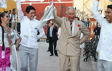 Prince Charles dancing with a group in traditional attire in Mexico, smiling and holding white handkerchiefs.