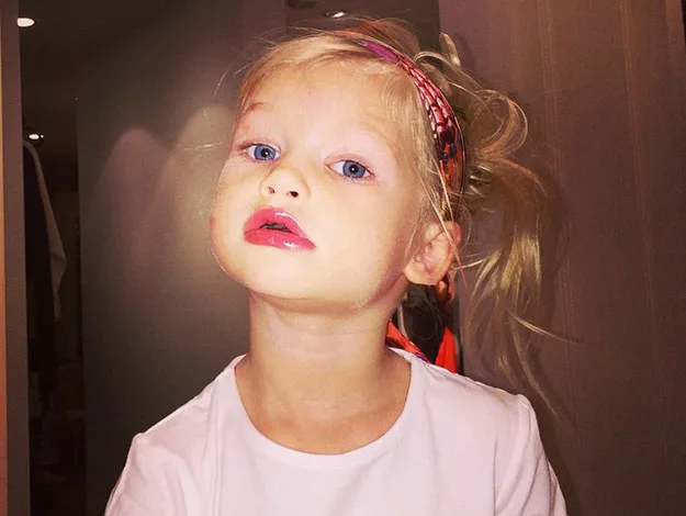 Young child with blue eyes and glossy lips, wearing a patterned headband and white shirt, looking upwards.