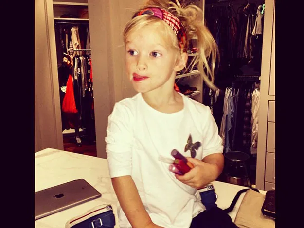 Young child with a headband, wearing a white shirt, holding makeup in a closet.