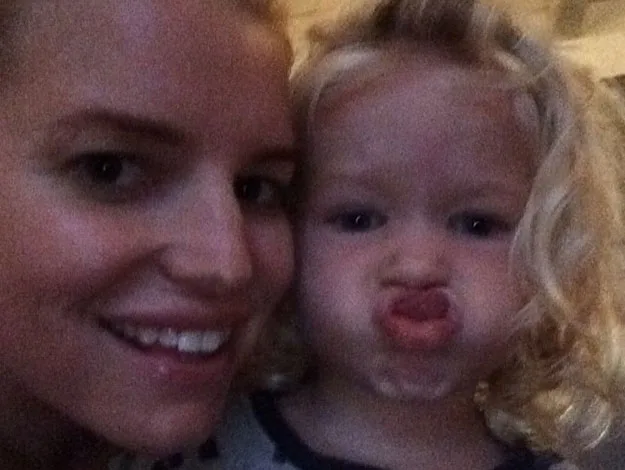 A woman smiles while a child with curly hair makes a funny face, pressing lips together, indoors.