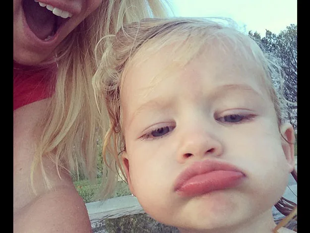 Toddler making a pouty face while posing for a selfie with an adult partially visible in the background smiling.