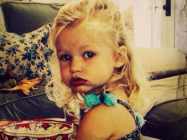A young child with curly blonde hair sitting in a living room, looking towards the camera with a serious expression.
